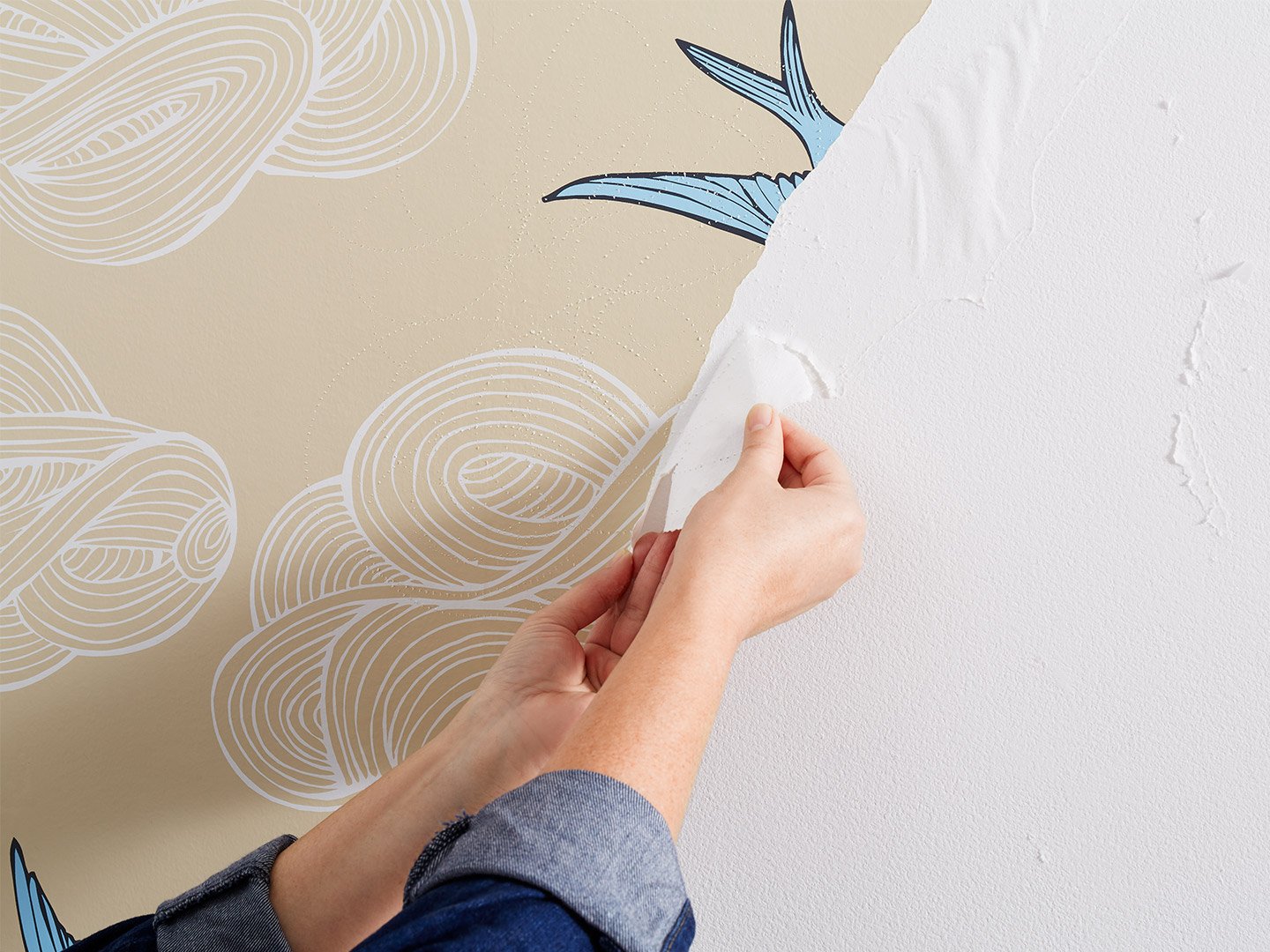 Two hands pulling beige wallpaper with white clouds and blue birds off a wall.